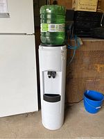 Full front view of the white water cooler with a large green water bottle on top, positioned next to a white fridge on a concrete floor.