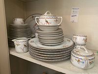 Shelf display of stacked porcelain dinnerware including large dinner plates, smaller dessert plates, saucers, teacups, and several covered serving pieces with bird and floral decoration