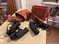 Two binoculars with leather cases on table, one case reddish with strap, other brown and open
