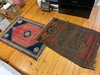 Photo showing two rugs on wooden floor: left carpet with blue, red, cream design; right worn kilim rug with geometric pattern in muted earth tones.