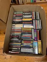 Top down view of a large cardboard box filled with stacked compact discs in their cases. Mostly classical music CDs observed.