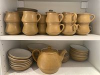 Middle shelf with a round mustard yellow stoneware teapot with lid, surrounded by stacked bowls and plates in matching color and style with beige interior.