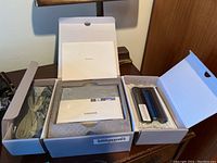 Open box showing Samsung photo printer accessories including power cable, USB cable, user manuals, and replacement cartridge.