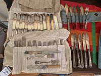 Full view of many knives and forks on cloth rolls and a colorful cloth, showing variety and number of flatware pieces.