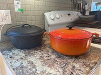 Two cast iron pots on kitchen counter: one raw cast iron pot with lid, one orange enamel Le Creuset Dutch oven with lid.
