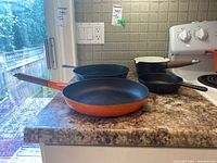 Photo showing the Le Creuset orange cast iron pan, a black enamel Copco pot, and two additional black cast iron pans on a countertop.
