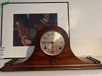 Full view of the wooden case mantle clock with curved top and scroll details on base set against a colorful framed background.