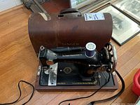 Vintage Singer portable electric sewing machine with attached power cord and spool of thread on wooden base next to original brown carrying case showing Singer branding.