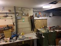 Wide view of workbench showing various tools including a blue drill, handsaw, metal axes, green multi-drawer cabinet, some small hardware containers and spray cans on window ledge.