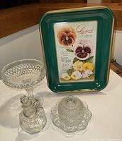 Green Burpee metal tray with pansy flower seed advertisement from 1910, shown standing behind glass items.