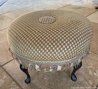 Front angle view showing the checkered fabric upholstery with centered button, fringe decorative trim, and curved black metal legs.