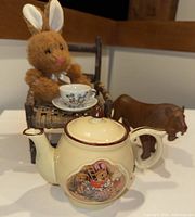 Photo showing the plush bunny in a twig chair holding the mini teacup and saucer, ceramic teapot and wooden lion figurine placed nearby on a flat surface.