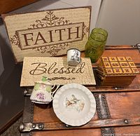 Overview of lot showing Faith and Blessed signs on wooden plaques, green embossed glass jar, enamelware rabbit cup, decorative plate, and porcelain floral container on wooden chest.