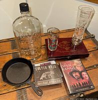Full lot arranged on wood surface showing all items including Bulleit bottle, Jim Beam skillet, shot glass, cigar box, CDs and DVD, beer glass.