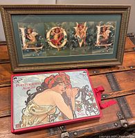 Photo showing the Mucha-themed photograph album with floral woman cover and the framed angel art spelling LOVE, placed on a wooden trunk.