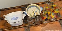 Overview of lot items on wooden surface showing Corning dishes, fruit juice glasses, metal shakers, and Quimper bowl