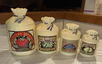 Full set of four ceramic canisters shaped and styled like burlap bags with produce labels, arranged on a table.