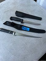 Photo showing two filet knives laid out on a surface, one with black plastic handle and black plastic sheath, the other with a black and metallic handle and fabric sheath with a strap.