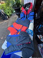 Four life jackets and a buoy displayed on a white table outside. Three life jackets are visible in different colors and styles. A white buoy with blue rope is also present.