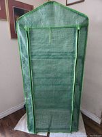 Fully assembled green mesh greenhouse with zippered front door, green trim, and peaked roof shape next to a wall.