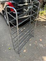 Front side of a gray metal shoe rack with silver frame set outdoors on pavement beside a vehicle.