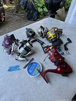 Lot of six fishing reels with two spare handles and spool of fishing line on outdoor table.