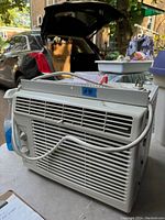 Photo showing front and side vent grills and control knobs of the white window air conditioner with power cord wrapped on top.