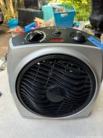 Front view of a gray Bionaire electric heater showing black round grille with fan behind, two control knobs on top, and a red indicator light. Label visible showing brand name.