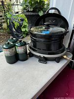 Coleman cooking station with removable black pot and glass lid accompanied by three green propane cans.