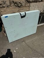 Table folded and standing vertically showing the handle and exterior surface with some marks and scuffs.