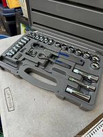 Open gray carrying case showing socket set including two ratchet handles with blue grips, multiple socket heads, extension bars, and adapters.