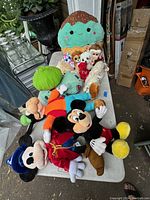 Photo of a variety of premium plush toys including Disney Mickey Mouse, Pokemon Bulbasaur, Ty Beanie Babies, and large green strawberry Squishmallow on a white table.