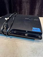 Photo of a black Sony PS3 console showing the top and front with a power cable.