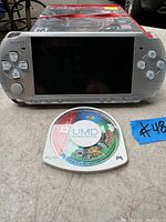 Front view of silver Sony PSP handheld console showing screen and buttons with UMD game disc placed in front.