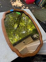 Photo showing an irregular shaped wall mirror with an oak wood frame placed on a light-colored surface, reflecting outdoor foliage and sky.