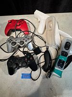 Lot of various gaming controllers placed on a table, showing Xbox One controller, multiple Wii controllers with wrist straps, and Sony PlayStation controllers (black and silver). Wired cables are visible.