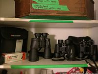 Shelf showing three pairs of binoculars including the large Celestron Skylux 15x70, Bushnell 8x40, and Fritz Hauff 7x35 with their respective carry cases and the tripod adapter box