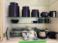 Blue ceramic canisters, mugs, tea cups, and dinnerware including plates and bowls inside a cabinet