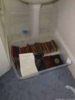 Plastic under bed storage tote containing approximately 20 vintage small books arranged lying flat with spines facing up and some accessories.
