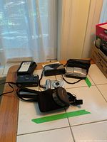 Lot of cameras and cases on a tiled floor including Kodak KB18 35mm camera, Canon PowerShot A580, Olympus camera and cases.