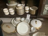 Stacked dinner plates, luncheon plates, bread and butter plates, saucers and tea cups on cabinet shelf with two lidded serving bowls below