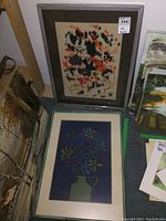 Two framed needlepoint artworks side by side, one floral vase and one abstract design, showing full frames and matting.