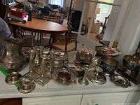 Wide shot of all silver plated items on a white counter including pitchers, candle holders, trays, coasters, and pedestal dish.