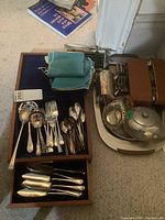Photo showing assorted silverplate cutlery in wooden canteen with blue felt lining, additional pieces stored in drawer, next to metal trays, pewter teapot, and brown leather travel vanity set with broken zipper.