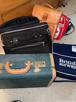 Group photo showing vintage suitcase, British Airways travel bag, brown leather satchel, black computer bag, and partially visible bags on carpeted floor