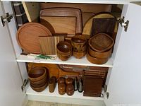Top shelf containing various wooden bowls, round and rectangular cutting boards, one striped square cutting board, and some trivets or small trays.