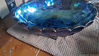 Blue carnival glass dish on a cloth with a wooden table underneath, showing the overall shape, scalloped edges and iridescent blue glaze.