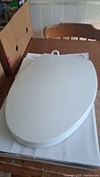 Photo of the closed white plastic toilet seat on its original box, showing smooth oval top surface and edges.