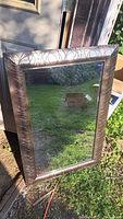 Full view of a rectangular wall mirror with metallic textured frame resting against a door, outdoors on grass.