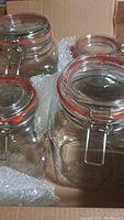 Four clear glass cannisters with metal snap-top clasps and orange sealing rings, positioned in a cardboard box with some bubble wrap.